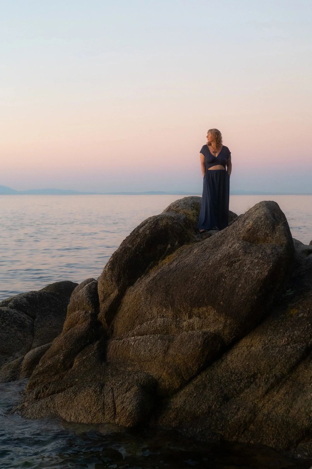 Juliana Christine in a navy dress standing on rocks by the water at sunset