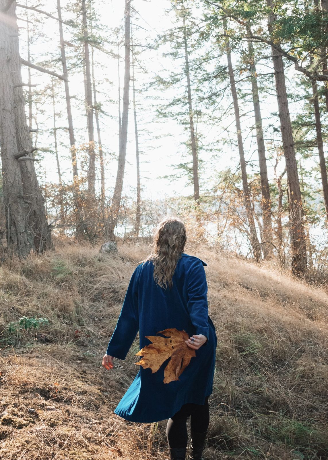 Juliana Christine in a blue coat walking through a forest, holding a large autumn leaf