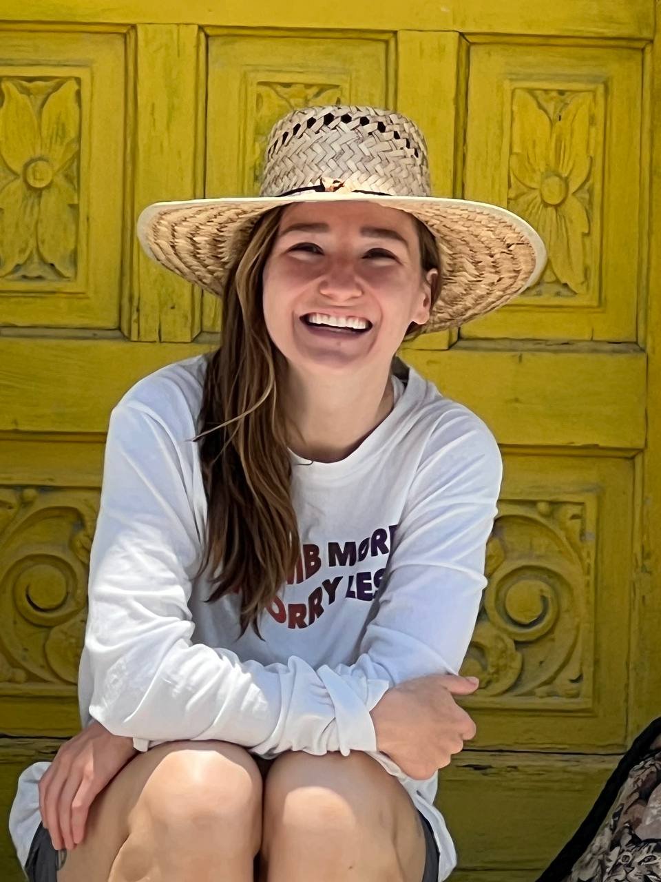 A smiling woman in a wide-brimmed straw hat and white shirt, sitting in front of a carved yellow wooden background