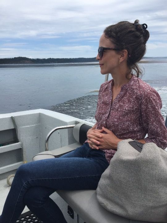A woman in sunglasses sitting on a boat with her hands in her lap, water and an island behind her